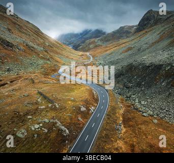 Luftaufnahme der gewundenen Bergstraße im Alpental bei bewölktem Herbsttag in der Schweiz. Blick auf die leere Straße im schweizer Bergpass, orange Stockfoto