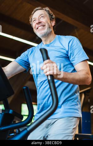 Ein älterer Mann lächelt, während er ein Heimtrainer fährt. Er trägt ein blaues Hemd und eine graue Hose Stockfoto