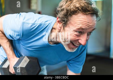 Nahaufnahme eines Reifen Mannes, der eine schwere Hantel hebt und lächelt Er ist im Fitnessstudio und trainiert seine Rückenmuskeln Er trägt ein blaues Hemd Stockfoto