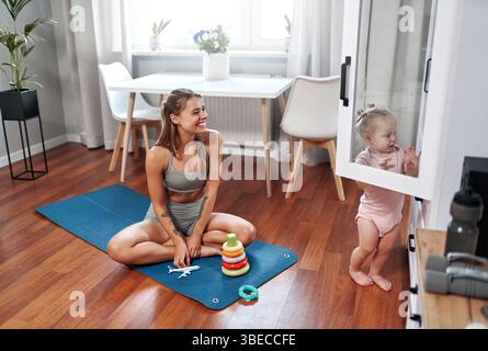 Eine fröhliche und fröhliche Mutter übt ihre Yoga-Routine zu Hause, während ihr liebenswertes Baby in der Nähe spielt und eine wunderbare Mischung aus Passform bietet Stockfoto