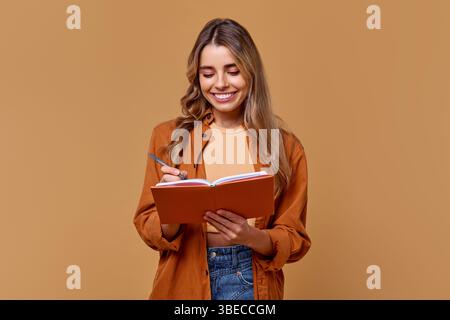 Eine junge Frau, die vor Freude strahlt und ein helles Lächeln auf ihrem Gesicht hat, ist tief in das Schreiben eingetaucht, fleißig in ihrem Notizbuch, vor einem warmen und unfreundlichen Hintergrund Stockfoto