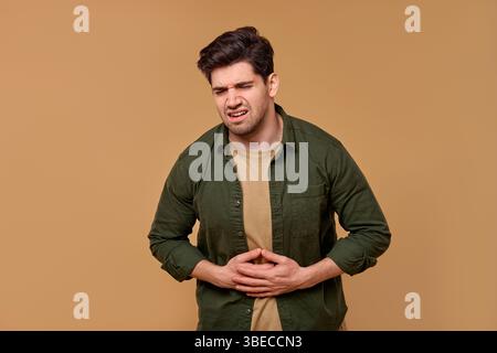 Ein junger Mann drückt sichtbares Unbehagen aus, während er seinen Magen hält, was in einer neutralen Umgebung festgehalten wird, die seine Gefühle nicht ablenkt Stockfoto