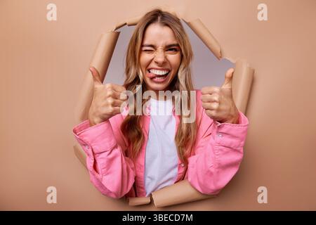 Eine fröhliche und überschwängliche junge Frau bricht durch eine lebendige Papierwand, ihr breites Lächeln strahlt positiv aus, während sie einen doppelten Daumen hochlegt, perfekt Stockfoto