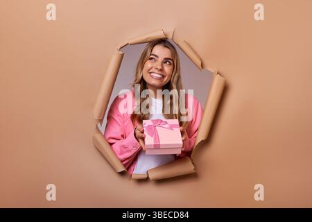 Eine fröhliche, lebendige junge Frau in einer entzückenden rosa Strickjacke hält ein ordentlich gewickeltes Geschenk und blickt aufgeregt durch ein Loch in warmem Braun Stockfoto