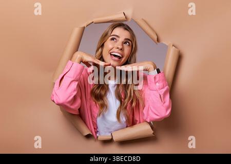 Eine fröhliche und fröhliche junge Frau mit schönen lockigen Haaren posiert spielerisch vor einem zerrissenen Papierhintergrund und strahlt ein wunderbares Gefühl von Glück aus Stockfoto