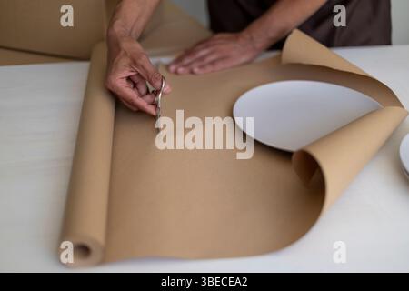 Person, die braunes Papier zum Einwickeln von Platten schneidet, wobei der Schwerpunkt auf Händen und Materialien liegt. Stockfoto
