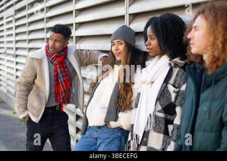 Vier verschiedene junge Studenten in Winterkleidung lehnen sich an eine Metallwand und genießen ihre gemeinsame Freizeit Stockfoto