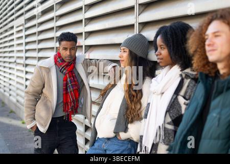 Vier verschiedene College-Studenten in Winterkleidung, die sich an eine Wand lehnen und zwischen den Unterrichtseinheiten eine Pause einlegen Stockfoto