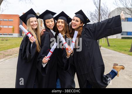Vier fröhliche, multiethnische Absolventen in akademischen Gewändern mit Diplomen, die ihren Abschluss außerhalb der Universität feiern Stockfoto