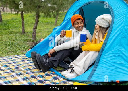 Junge Multi-ethnische Paare campen, trinken heiße Getränke in ihrem Zelt und genießen den Outdoor-Lebensstil Stockfoto
