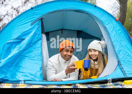 Das junge, ethnische Paar lächelt und genießt warme Getränke beim Camping in der freien Natur und genießt den gemütlichen Komfort der Natur Stockfoto