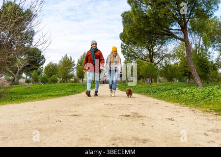 Junge Paare, die mit ihrem verspielten Dackel durch einen Park schlendern, sich in der Wärme eines sonnigen Tages erfreuen und die Natur genießen Stockfoto