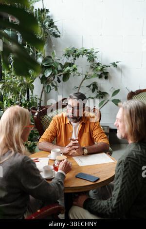 Eine Gruppe von drei Personen, die an einem runden Tisch sitzen und Pläne in gemütlicher Gartenatmosphäre diskutieren, umgeben von üppigen grünen Pflanzen, sieht entspannt aus und führt Gespräche Stockfoto