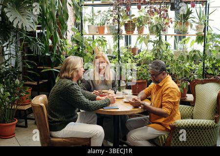 Drei Personen genießen Kaffee an einem Holztisch in einer üppigen grünen Umgebung mit vielen Pflanzen. Tageslicht, das durch große Fenster gefiltert wird, unterstreicht die gemütliche Atmosphäre Stockfoto