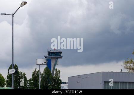 Nürnberg, Deutschland - 25. Mai 2025: Flughafenkontrollturm unter bewölktem Himmel. Straßenlaternen und Laub im Vordergrund. Gebäude auf der rechten Seite. Stockfoto