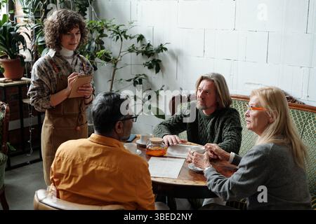 Gruppe von Kollegen, die sich um einen Holztisch in gemütlicher Büroumgebung mit Pflanzen und natürlicher Einrichtung unterhalten, die Wärme und Komfort verleihen Stockfoto