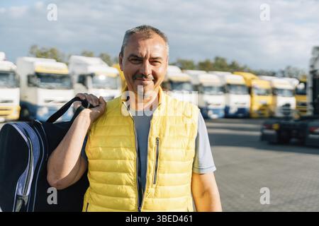Porträt eines kaukasischen Reifen Mannes mit Tasche auf Parkhintergrund einiger LKW-Fahrzeuge. Lkw-Fahrer-Arbeiter. Stockfoto