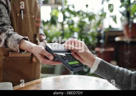 Person, die für Kaffee mit kontaktloser Karte in einem lokalen Café bezahlt. Barista hält Zahlungsterminal mit grünen Pflanzen im Hintergrund Stockfoto
