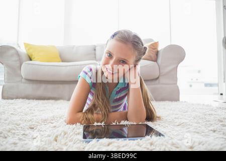 Schulmädchen, das auf einem weißen Teppich im hellen Wohnzimmer liegt und auf die Tablette blickt Stockfoto