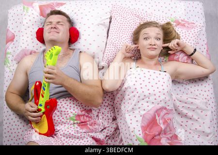 Junges Paar im Bett liegend. Mann spielt Gitarre und Schreie seiner Stimme. Frau bedeckt ihre Finger in die Ohren Stockfoto
