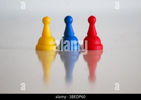 Drei Spielfiguren in Gelb, Blau und Rot, die den Flaggen Kolumbiens, Venezuelas oder Ecuadors ähneln, auf einer reflektierenden weißen Oberfläche. Stockfoto