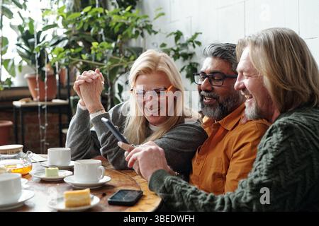 Eine Gruppe von Freunden genießt Kaffee und Kuchen, während sie sich unterhalten und lachen in einem gemütlichen Café mit Pflanzendekor und natürlichem Licht Stockfoto