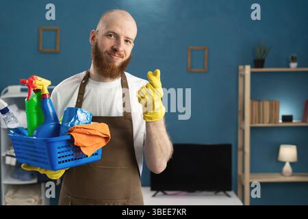 Glücklicher Hausmeister, bärtiger Mann, der einen Korb mit Putzmitteln hält, zeigt Liebesherz-Geste. Reinigungs-, Housekeeping- und Housework-Konzept Stockfoto
