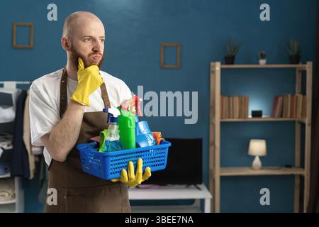 Hübscher bärtiger Glatzkopf, der Reinigungsprodukte hält und nachdenklich sein Kinn kratzt, während er wegblickt und an einen Plan im Wohnzimmer denkt. Househ Stockfoto