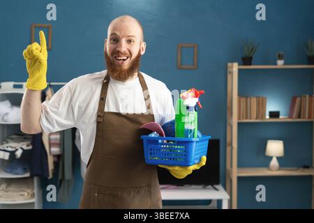Glücklicher bärtiger Mann in Schürze, der einen Korb mit Putzmitteln hält, lächelt in die Kamera und zeigt Daumen hoch, Geste der überlegten Idee, Genie. Kopierbereich Stockfoto