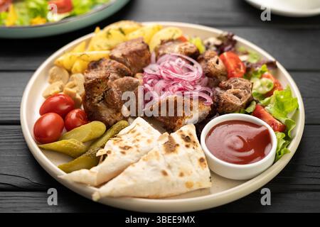 Gegrillter Fleischkebab, serviert mit Lavash gefüllt mit Suluguni-Käse, Kirschtomaten, gebratenen Kartoffeln, eingelegten Gurken, Ketchup und Zwiebeln auf Teller Stockfoto