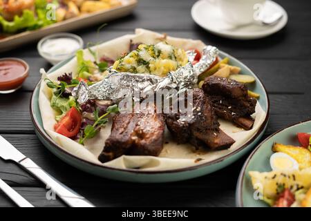 Nahaufnahme gegrillter Schweinerippchen mit gebackenen Kartoffeln und Gemüse auf dem Esstisch im Restaurant, Konzept von gegrilltem Fleisch und herzhafter Mahlzeit Stockfoto