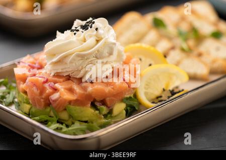 Frischer Lachstartar mit Avocado, roten Zwiebeln und Rucola, garniert mit Sahne und schwarzen Sesamsamen, serviert mit Zitronenscheiben und geröstetem Brot, gourm Stockfoto