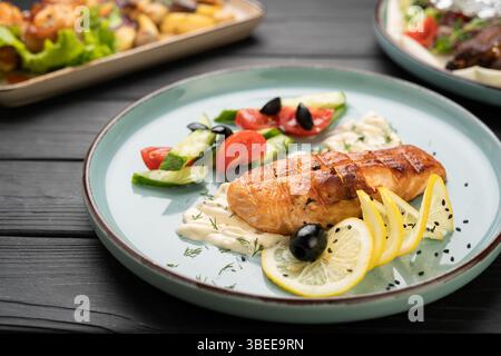 Gegrilltes Lachsfilet serviert mit cremiger Sauce, frischem Gemüse und Zitronenscheiben auf Teller im Restaurant, Meeresfrüchte-Rezept für das Restaurant-Menü pro Person Stockfoto