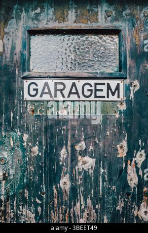 Verwittertes Vintage Garagentor mit abblätternder grüner Farbe zeigt portugiesisches Garagem-Schild auf einer versehrten Holzoberfläche. Rustikale Küstenarchitektur Stockfoto