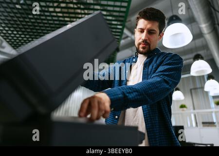 Junger Mann, der einen modernen Bürodrucker bedient, während er sich auf das Drucken von Dokumenten an einem ruhigen Arbeitsplatz konzentriert Stockfoto