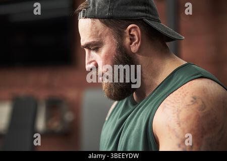 Bärtiger Mann mit ärmellosem Hemd und rückwärtiger Mütze, verlor während des Fitnessstudiums Gedanken. Sichtbare Tattoos auf Arm und Fitnessgeräte im Hintergrund Stockfoto