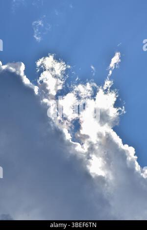 Helle Sonnenstrahlen durchbrechen weiße Wolken an einem klaren blauen Himmel, natürliches Licht und friedliche Atmosphäre. Stockfoto