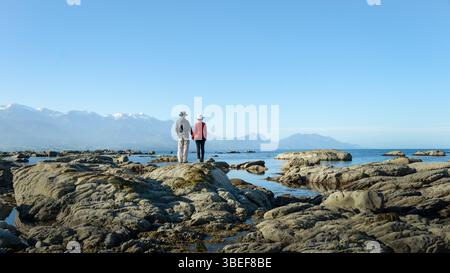 Pärchen stehen am steinigen Strand. Schneebedeckte Berge in der Ferne. Kaikoura. Südinsel. Stockfoto