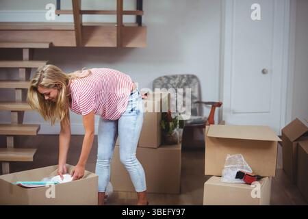 Frau, die sich über Pappkarton beugt und Verpackungsmaterialien mit rotem Klebeband im Wohnzimmer auspackt Stockfoto