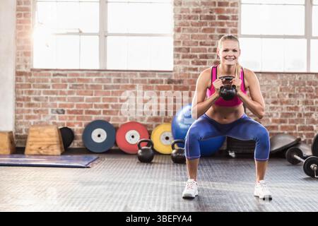 Frau, die eine Kettlebell auf der Brust hält, während sie im Fitnessstudio mit farbigen Hantelplatten hockt Stockfoto