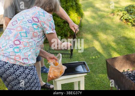 Älteres älteres Ehepaar, das sich bereit macht, marinierte Speisen am Sommertag im Garten zu grillen. Pensionierter Mann und Frau bereiten draußen Grillfest vor. Stockfoto