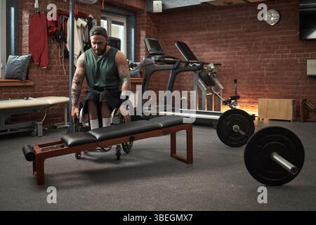 Mann mit Rollstuhl, der im Fitnessstudio mit Gewichtheben und Cardio-Geräten im Hintergrund zu sehen ist. Rollstuhlsportler, der Fitness und Strapazierfähigkeit aufrechterhält Stockfoto
