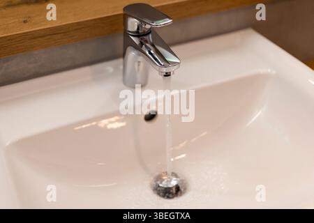 Ein eleganter Chromhahn liefert einen gleichmäßigen Strahl von klarem Wasser in ein sauberes, weißes Waschbecken. Das moderne Badezimmer verfügt über warme Holzakzente und einen Stockfoto
