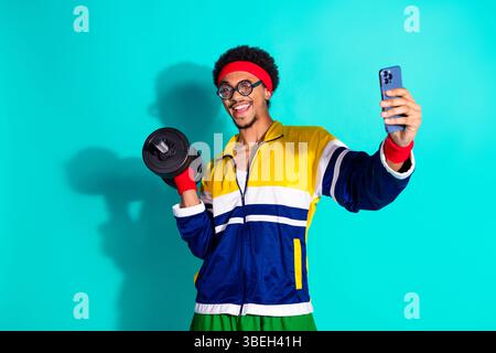 Junger Mann in Retro-Sportswear lächelt, während er die Kurzhantel hält und ein Smartphone-Selfie auf einem leuchtenden blaugrünen Studiohintergrund macht Stockfoto