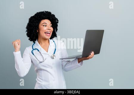 Fröhliche Ärztin feiert Leistung, während sie einen Laptop hält, vor grauem Hintergrund in einer medizinischen Uniform steht Stockfoto