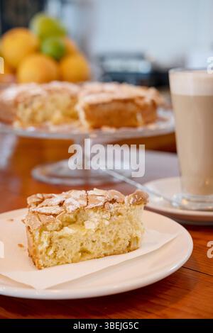 Ein frisch geschnittenes Stück hausgemachter Mandel- und Apfelkuchen auf einem Teller mit Serviette, ein großer Latte in einem hohen Glas und ein ganzer Kuchen auf einem Glasständer Stockfoto