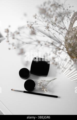 Minimalistischer Beauty Flat Lay mit Black Cosmetics and Flowers Ein flaches Lay von Schönheitswerkzeugen, einschließlich schwarzer und rosa Mascara-Sträuße, mit zartem weißen flo Stockfoto