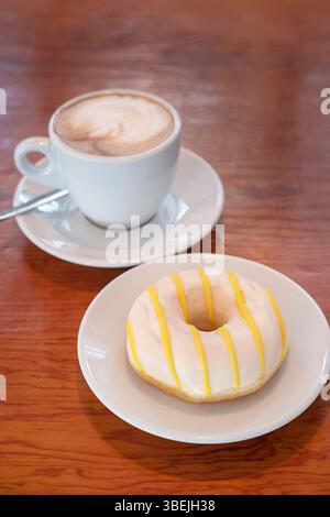 Cappuccino aus Keramik mit Latte Art, serviert auf einer Untertasse mit einem Löffel, begleitet von einem weiß-mattierten Krapfen mit hellgelber Zuckerguss Stockfoto