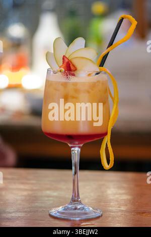 Tequila Sunrise, ein lebendiger alkoholischer Cocktail in einem hohen Stielglas, garniert mit frischen Apfelscheiben, einem Erdbeerkeil und einer Orangenschale Stockfoto