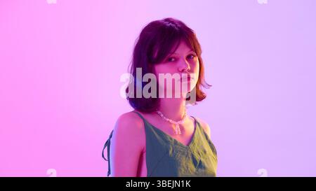 Stilvolles Teenager-Mädchen mit Piercings in grünem Kleid und Perlenkette, das ruhig unter lila rosa Beleuchtung vor verlaufendem Hintergrund aussieht Stockfoto